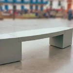 Banca de concreto moderna sin espaldar, instalada en una plaza pública de un pueblo colombiano, con personas y edificios coloniales al fondo.