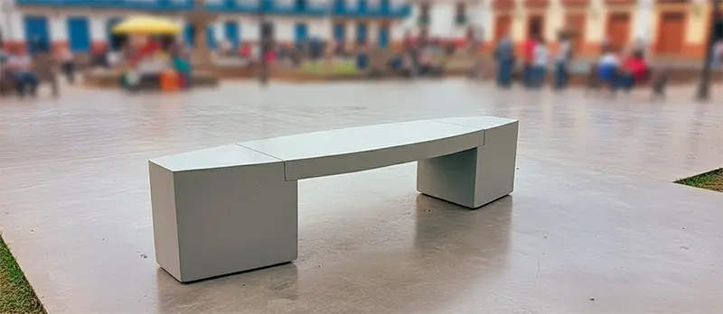 Banca de concreto moderna sin espaldar, instalada en una plaza pública de un pueblo colombiano, con personas y edificios coloniales al fondo.