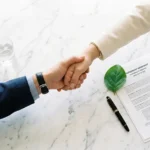 El aliado ideal para la Ley 2173: 5 claves para potenciar su impacto ambiental 1 Fotografía minimalista y elegante de un apretón de manos corporativo sobre una mesa de juntas, junto a una hoja verde nativa y un documento legal. Representa la alianza estratégica y la confianza B2B entre las empresas de Bogotá y Vivero Harmony, el operador forestal experto elegido para garantizar el cumplimiento de la Ley 2173 (Ley del Árbol) mediante proyectos de reforestación llave en mano